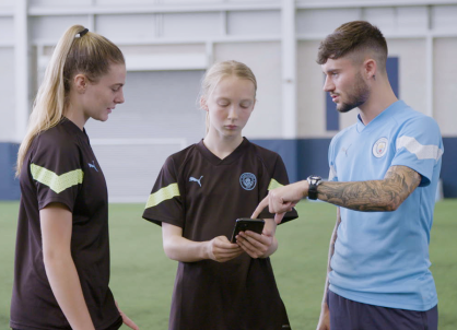 A Manchester City coach explaining to 2 female footballers how the CITYPLAY app works